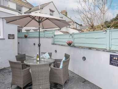 An outdoor patio with a round glass table and six wicker chairs under a large umbrella at Cobblers Cottage in Beaumaris