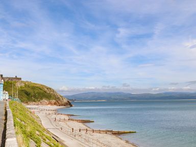 A coastal promenade with a pathway along the beach and a hillside with buildings near Menuchah in Criccieth