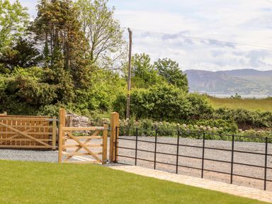 A grass lawn with a wooden gate and metal fence with trees and hills in the background at The Stables in Penmon near Beaumaris