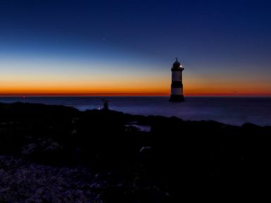 A lighthouse in the sea with a person on the rocky shore at dusk at The Stables in Penmon near Beaumaris