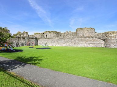 A green field with playground equipment and pathway next to a stone castle wall at The Stables in Penmon near Beaumaris