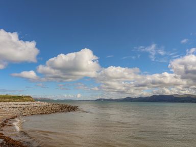 A rocky shoreline with water and distant hills under a blue sky with scattered clouds at The Stables in Penmon near Beaumaris