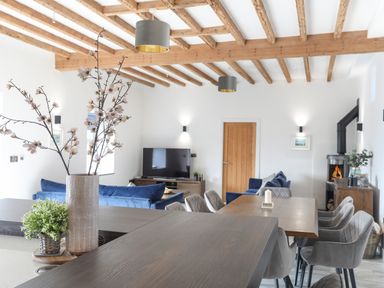 An open living and dining room with wooden beams on the ceiling a dining table with gray chairs a fireplace and blue sofas at The Stables Penmon near Beaumaris