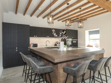 A kitchen with a large wooden island and six gray bar stools under wooden ceiling beams at The Stables in Penmon near Beaumaris