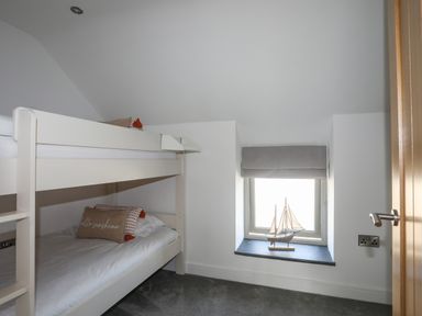 A small bedroom with white bunk beds and a window with a model sailboat on the sill at The Stables in Penmon near Beaumaris