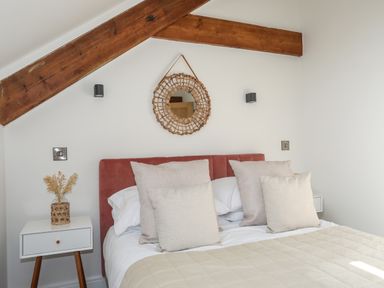 A bedroom with a bed featuring beige pillows and a beige quilt under wooden beams and a round woven mirror at The Stables in Penmon near Beaumaris