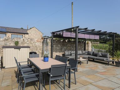 An outdoor patio with a dining table and chairs a sectional sofa under a pergola and a stone wall at The Stables in Penmon near Beaumaris