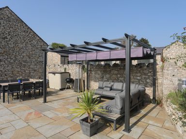 An outdoor patio with gray cushioned seating under a pergola a dining table with chairs a barbecue and a potted plant at The Stables in Penmon near Beaumaris