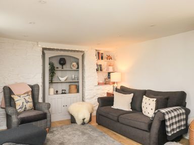 A living room with a dark sofa and gray armchair next to a white stone wall with shelves and a lamp at Hope Cottage in Buckfastleigh