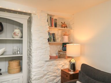 A corner with white stone walls featuring built-in shelves with books and decorative items a wooden side table with a lamp and a dark sofa at Hope Cottage in Buckfastleigh