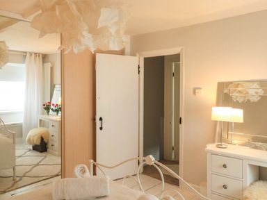 A bedroom with a white metal bed, rolled towels, a white dresser with a lamp and mirror, an open door, and a window with white curtains at Hope Cottage in Buckfastleigh