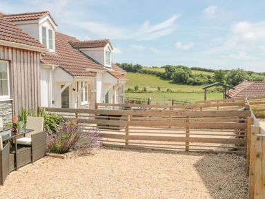 An outdoor patio area with a table and chairs beside a house with a wooden fence and green fields in the background at Garland in Nottington near Weymouth