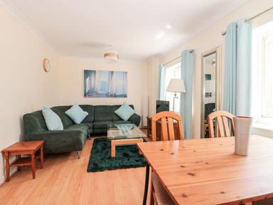 A living room with a green sofa blue cushions a glass coffee table wooden dining table and chairs near windows with blue curtains at Garland in Nottington near Weymouth