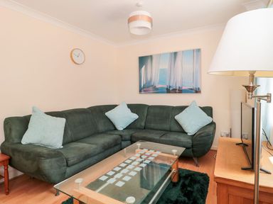 A living room with a dark green sectional sofa and light blue cushions a glass coffee table a wooden side table with a lamp a wall clock and a wall picture at Garland in Nottington near Weymouth
