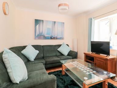 A living room with a green sofa and light blue cushions a glass coffee table a wooden TV stand with a television and a large window with curtains at Garland in Nottington near Weymouth