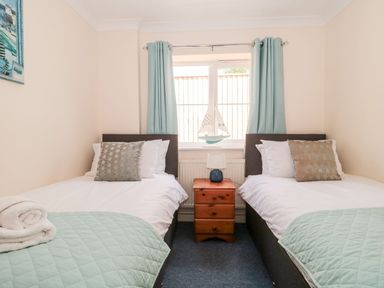 A bedroom with two single beds separated by a wooden bedside table with a lamp and a window with blue curtains and a decorative sailboat at Garland in Nottington near Weymouth