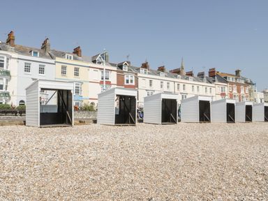 A row of small white beach huts on a pebble beach with colorful houses in the background at Garland in Nottington near Weymouth