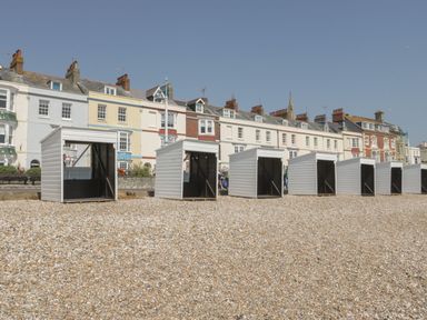 A row of small white beach huts on a pebble beach with colorful buildings in the background at Mellstock in Nottington near Weymouth