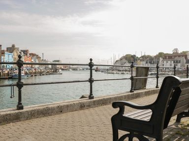 A waterfront scene with benches, railing, boats docked, and buildings along the water at Mellstock in Nottington near Weymouth