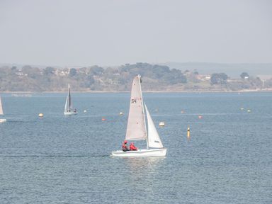 A sailboat with two people on a body of water with a shoreline and trees in the background at Mellstock in Nottington near Weymouth