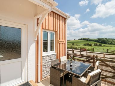 An outdoor patio area with a glass table and four wicker chairs next to a wooden building with a window and a sign reading Emminster with green fields and a wooden fence in the background at Emminster in Nottington near Weymouth