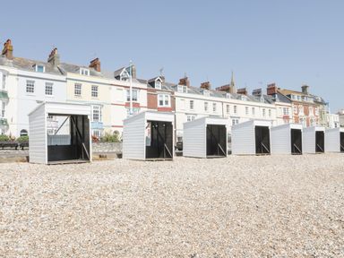 A row of white beach huts on a pebble beach with residential buildings in the background at Emminster in Nottington near Weymouth