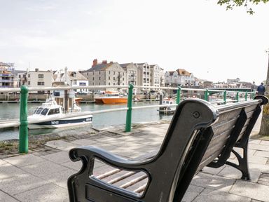 A black bench beside a waterfront with boats and buildings in the background at Emminster in Nottington near Weymouth