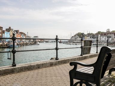A bench on a paved walkway next to a waterfront with boats and buildings at Emminster in Nottington near Weymouth