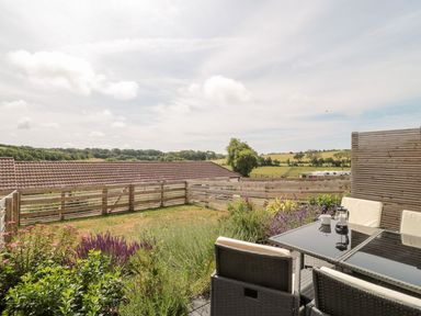 An outdoor patio with a glass table and cushioned chairs next to a garden with plants and a wooden fence at Egdon Heath in Nottington near Weymouth