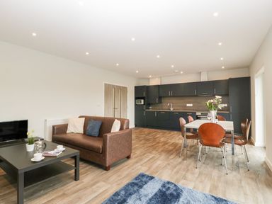 An open plan living room and kitchen with a brown sofa black coffee table dining table with chairs and kitchen cabinets at Dirdale in Weymouth