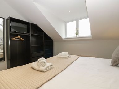 A bedroom with a bed covered by a beige quilt and two sets of folded towels a black shelving unit with a mounted television and hangers and a window with a plant on the sill at Dirdale in Weymouth