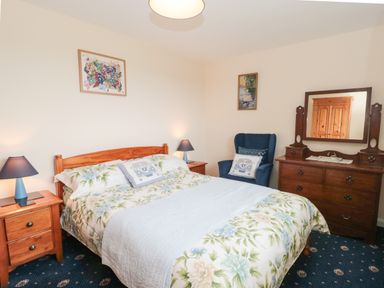 A bedroom with a bed, two bedside tables with lamps, a blue armchair, a wooden dresser with a mirror, and two framed pictures on the walls at Broadstones Bridport