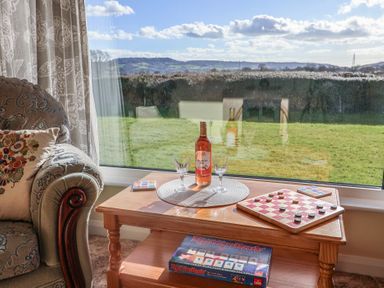 A wooden coffee table with a wine bottle glasses and a board game next to a patterned armchair and window at Broadstones in Bridport