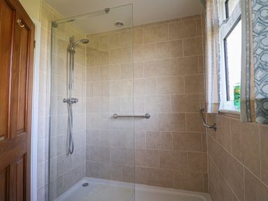 A tiled shower area with a glass screen a handheld showerhead and a window with blue patterned curtains at Broadstones in Bridport