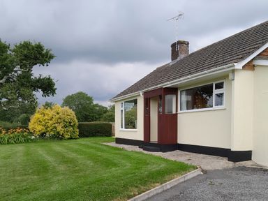 A single story house with a sloped roof near a lawn with bushes and trees at Broadstones in Bridport