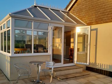 A conservatory with glass doors and windows opening to a patio with metal table and chairs at Broadstones in Bridport