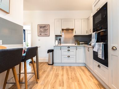 A kitchen with white cabinets wooden floor two dark gray bar stools and a trash can at Anchor House in Weymouth