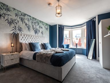 A bedroom with a large bed blue cushions bedside table with lamp bay window with blue curtains and a mirror on the floor at Anchor House in Weymouth