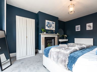 A bedroom with a bed draped in blue and gray covers a white wardrobe a fireplace with plants and a painting and a standing mirror at Anchor House in Weymouth