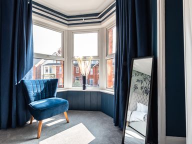 A corner with large windows a blue chair dark blue curtains a standing mirror and a vase with dried flowers at Anchor House in Weymouth