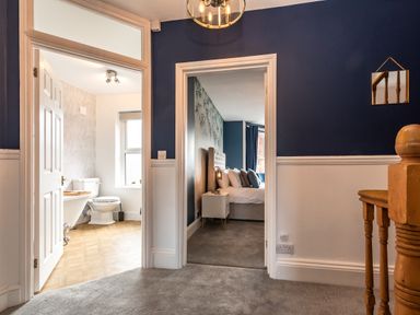 A hallway with a staircase bannister leading to a bathroom with a toilet and bathtub and a bedroom with a bed and bedside table at Anchor House in Weymouth