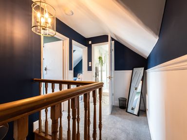 A hallway with blue and white walls wooden railing carpeted floor a hanging light fixture an open door leading to a bathroom and a leaning mirror at Anchor House Weymouth