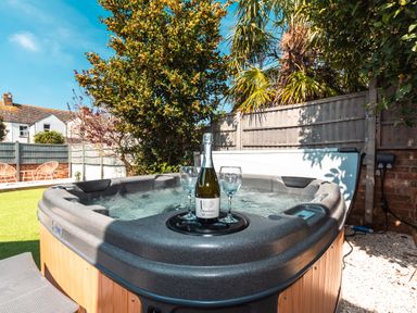 An outdoor hot tub with a bottle of prosecco and two glasses in a garden with trees and chairs at Anchor House in Weymouth