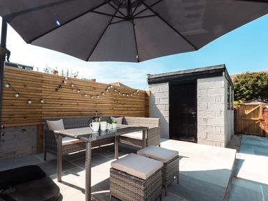 An outdoor patio with a rattan table and chairs under a large umbrella next to a wooden fence and a shed with a black door at Anchor House in Weymouth