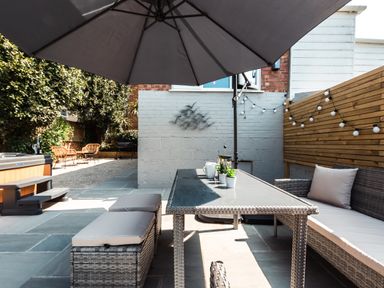 An outdoor patio with a glass table under a large umbrella a cushioned bench and stools string lights and a wooden fence at Anchor House in Weymouth
