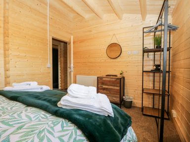 A bedroom with wooden walls a bed with green leaf patterned bedding and white towels a wooden dresser a round wall mirror and a black metal shelving unit at Howgills Retreat in Gatebeck near Kirkby Lonsdale