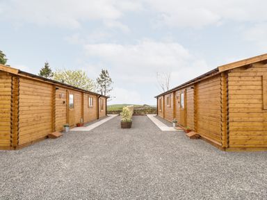 Two wooden cabins facing each other with gravel ground and a planted barrel in the center at Howgill Lodge in Gatebeck near Kirkby Lonsdale