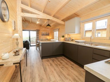 A kitchen with gray cabinets and wood flooring in a wooden lodge with a dining area and a living room visible at Howgill Lodge Gatebeck near Kirkby Lonsdale