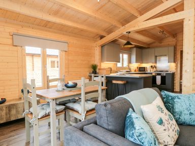 A dining area with a wooden table and chairs next to a kitchen with grey cabinets and a grey sofa in a wooden cabin at Howgill Lodge in Gatebeck near Kirkby Lonsdale