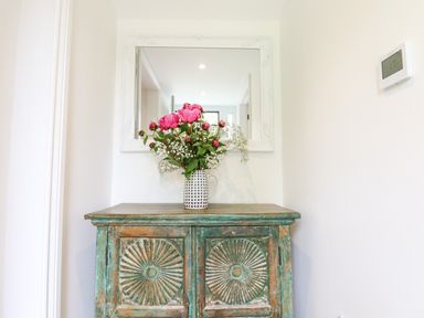 A distressed wooden cabinet with a vase of pink flowers and a white framed mirror above it at Bluebell Cottage in Mary Tavy